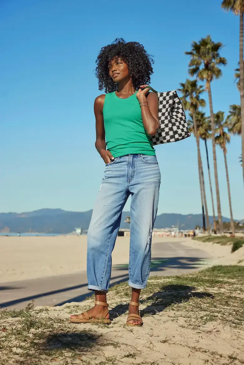 A woman with curly hair wearing a green sleeveless top and light blue jeans stands on a beach. She is carrying a black and white checkered bag over her shoulder and wearing tan sandals. Palm trees and mountains are visible in the background. This look features wardrobe staples, best basics, ideal for a capsule wardrobe.