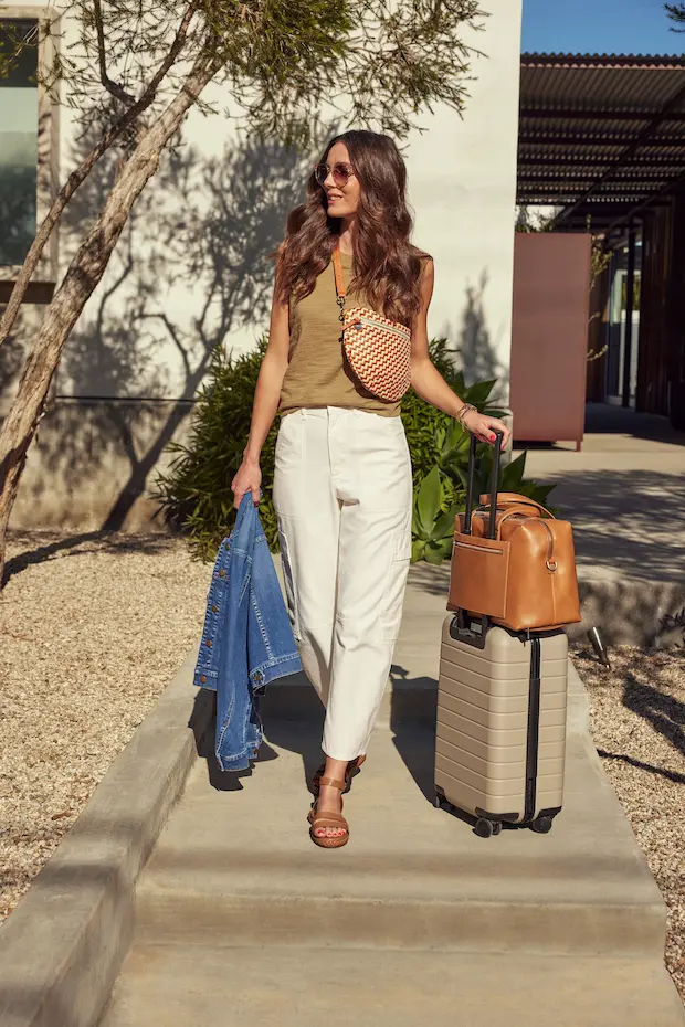 A woman with long brown hair wearing a green sleeveless top, white pants, and brown sandals is walking outside with a rolling suitcase and a tan handbag. She is also carrying a denim jacket and a woven crossbody bag. This casual outfit is perfect for what to wear on the weekend and is an essential part of a summer vacation packing list.